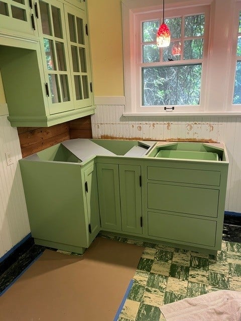 Green kitchen cabinets in renovation with open drawers, pink window frame, and vintage tile flooring